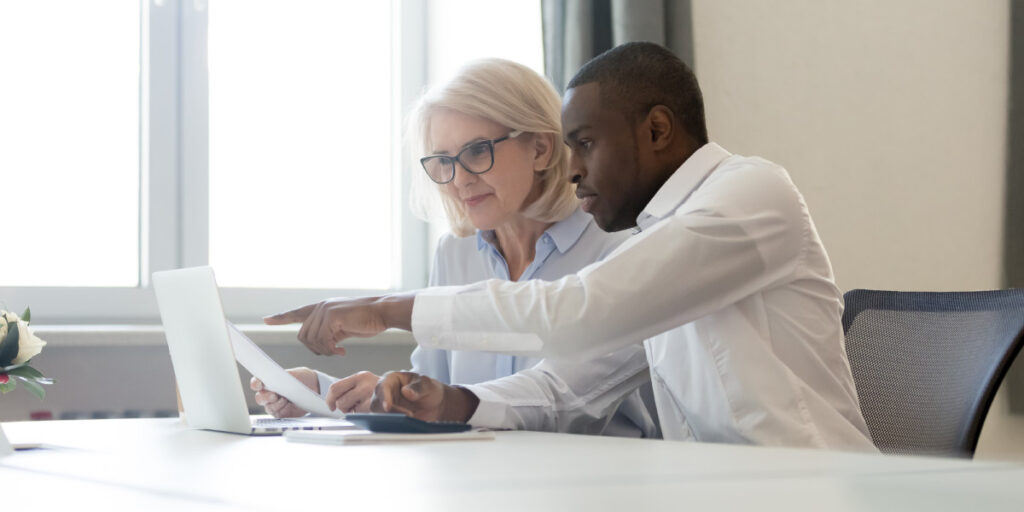 A young man assist a woman with reviewing a policy update printed and on a laptop.